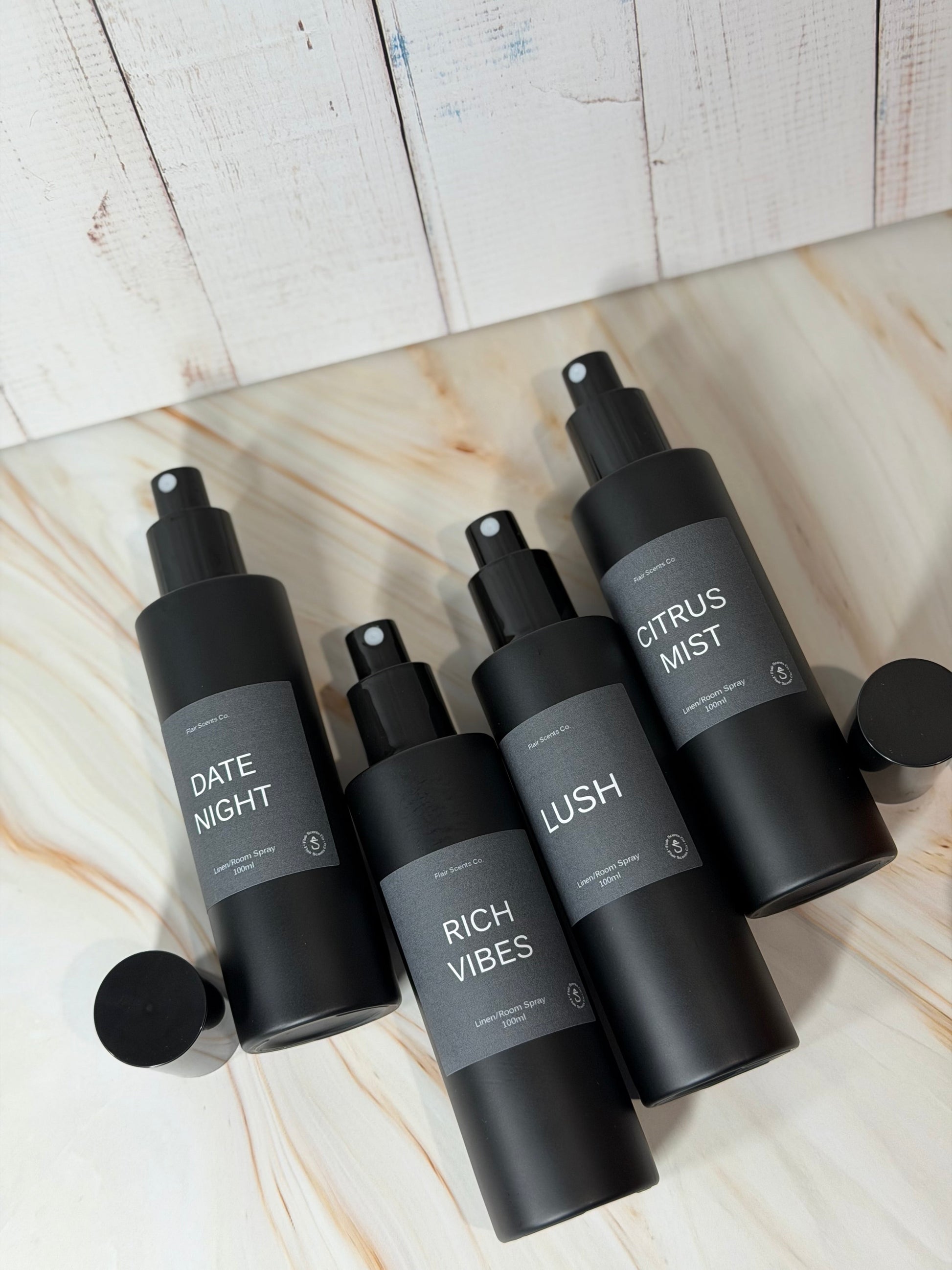 Set of four black matte Room/Linen Spray bottles with labels Date Night, Rich Vibes, Lush, and Citrus Mist on a marble surface.