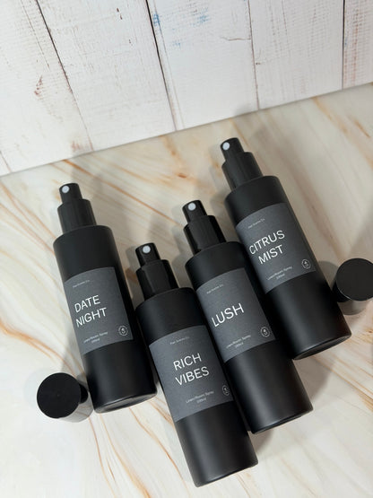 Set of four black matte Room/Linen Spray bottles with labels Date Night, Rich Vibes, Lush, and Citrus Mist on a marble surface.